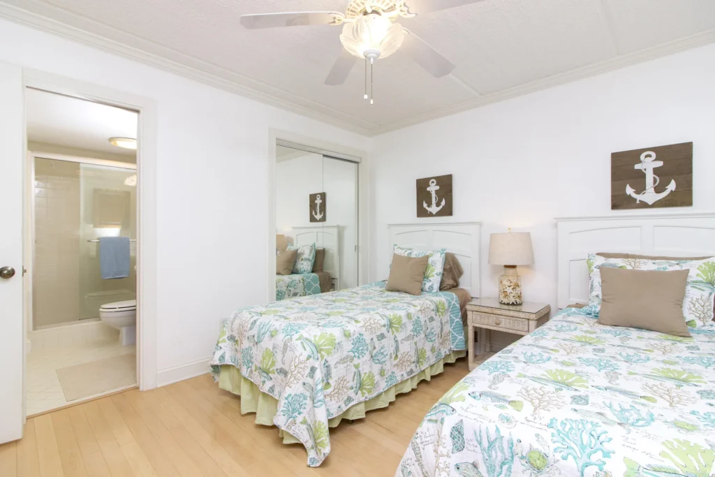 Two twin beds with sea-themed bedding flank a wicker nightstand and lamp, adjacent to a bathroom with glass shower doors.