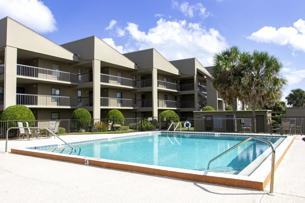 Rectangular pool with metal handrails, surrounded by a concrete deck and beige three-story condo building.