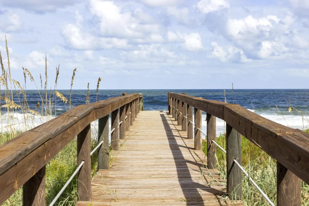 Wooden boardwalk with sturdy railings leads through dune grass to the ocean under a partly cloudy sky.