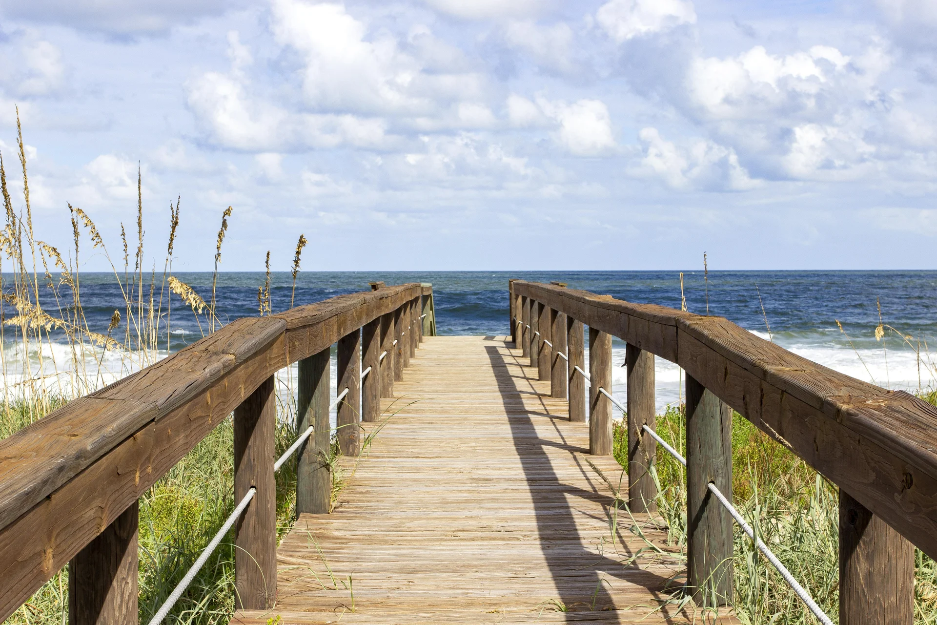 Wooden boardwalk with sturdy railings leads through dune grass to the ocean under a partly cloudy sky.