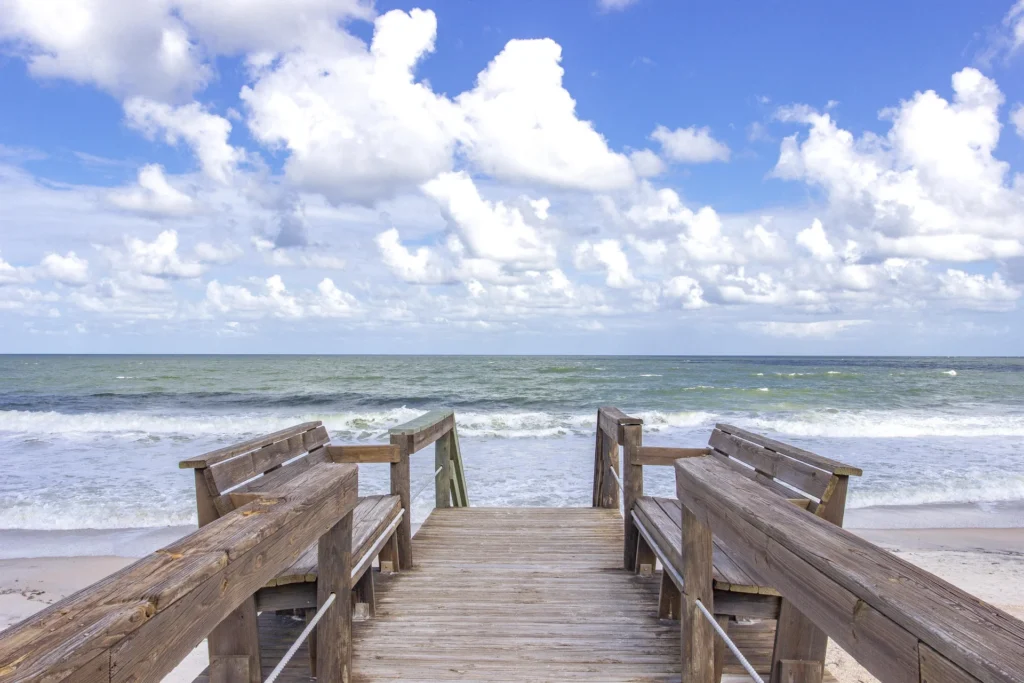 Wooden boardwalk with built-in benches leads down to sandy beach and gentle ocean waves under a partly cloudy sky.