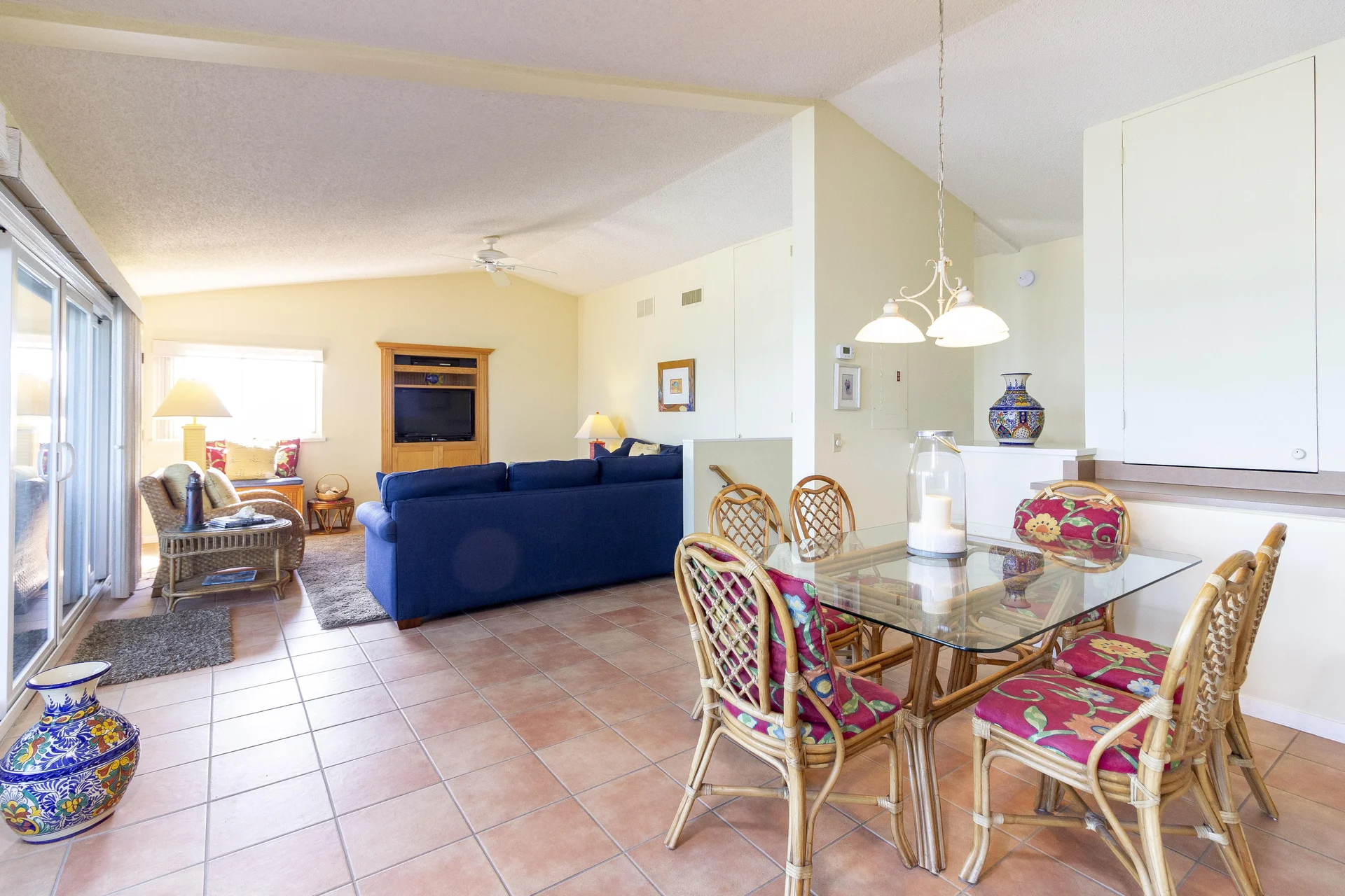Glass-top dining table with floral cushioned rattan chairs sits beside a tiled floor living area with blue sofas and wooden entertainment center.