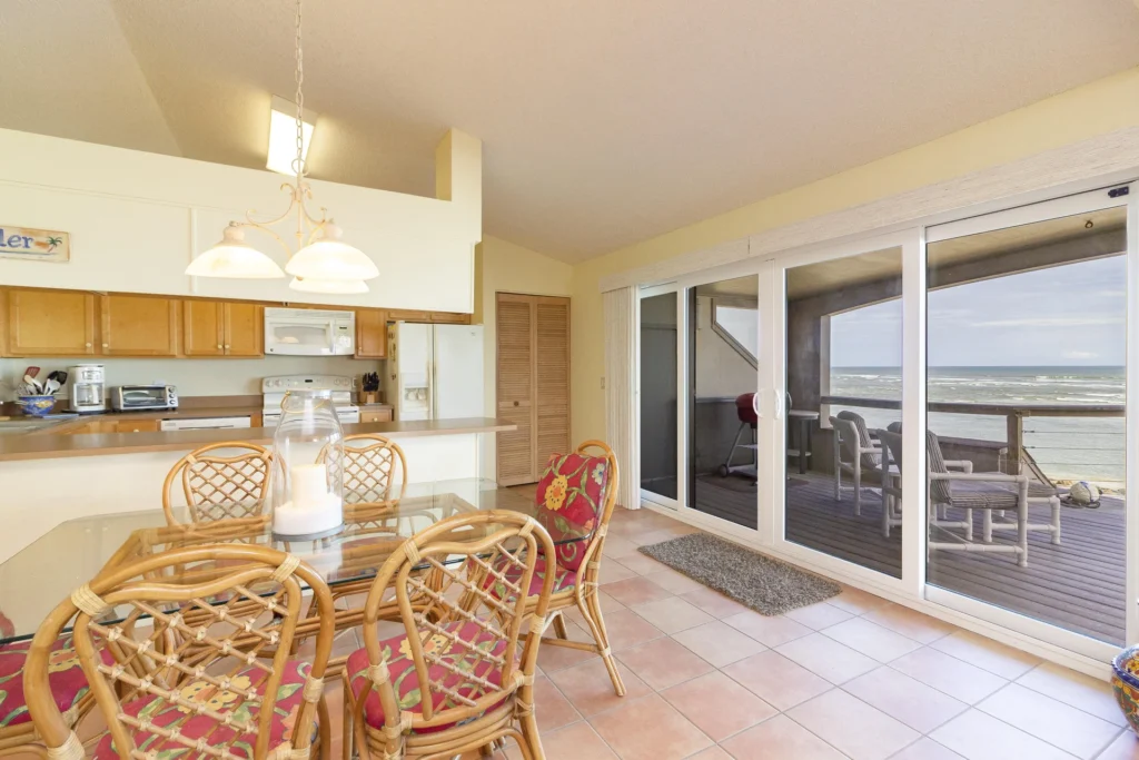 Dining area with glass-top table, wicker chairs, and sliding glass doors opening to oceanfront deck.