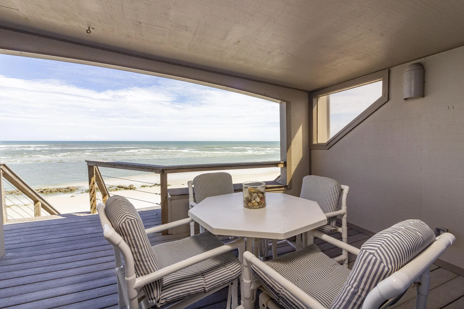 Covered balcony with white octagonal table, striped cushioned chairs, and oceanfront views over the beach.