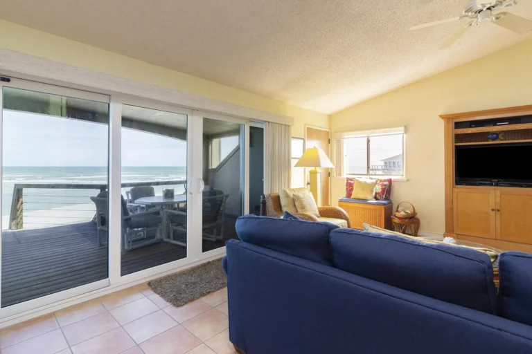 Living area with blue sofa, wicker armchair, wooden entertainment center, and sliding glass doors to oceanfront deck.