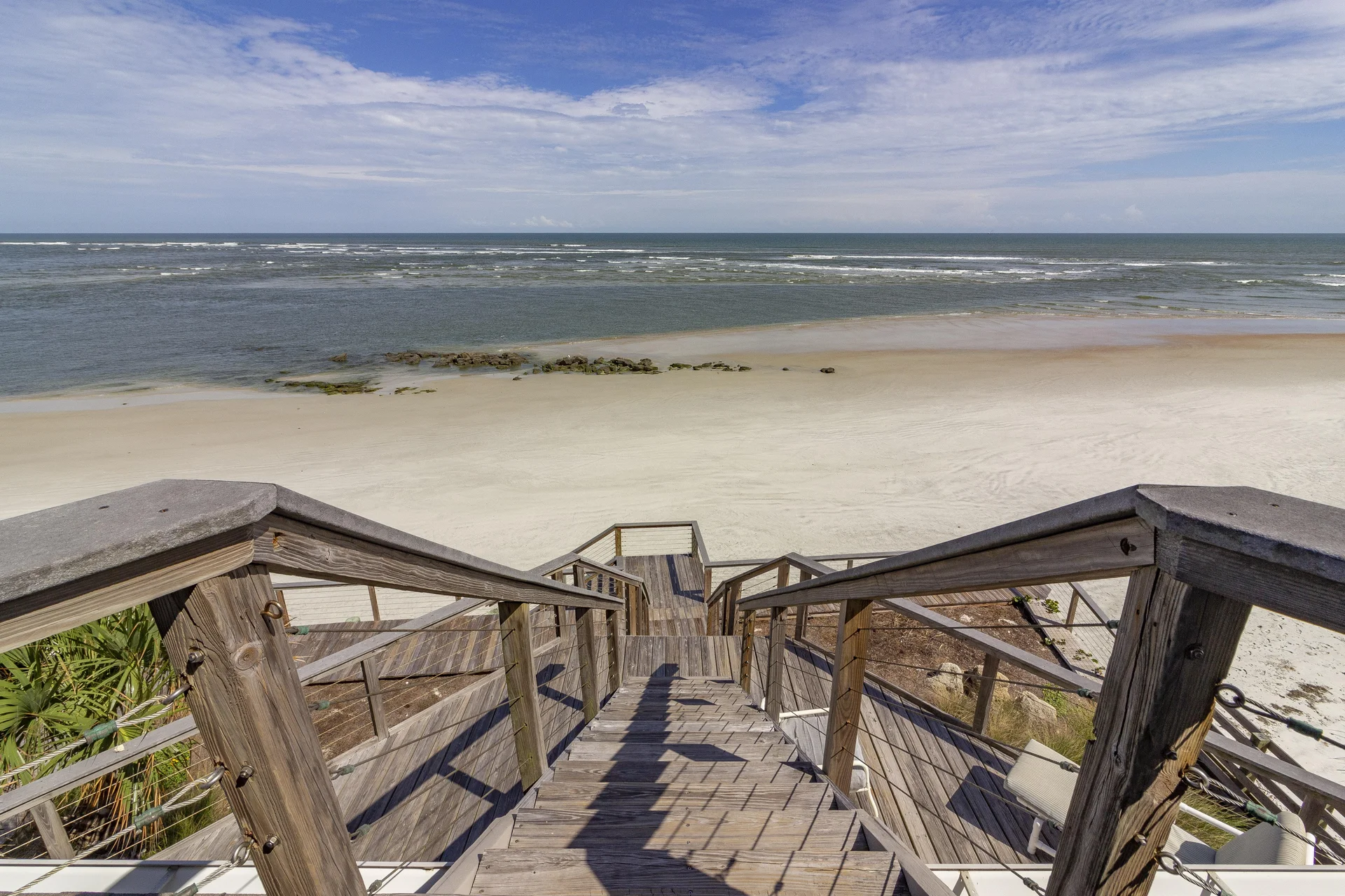 Sunlight casts shadows on the weathered wood steps and railing leading down to the sandy beach and ocean.