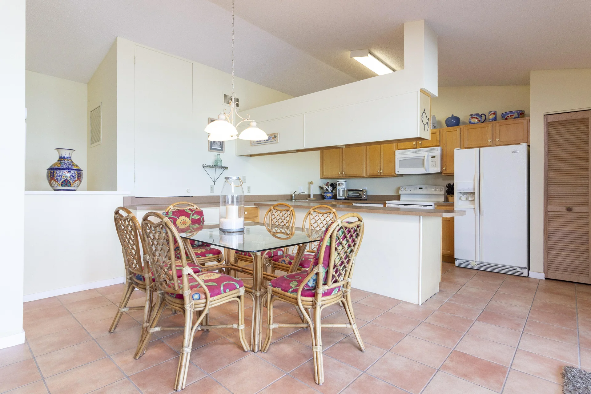 Glass-top dining table surrounded by six rattan chairs with red floral cushions on terracotta tile floor.