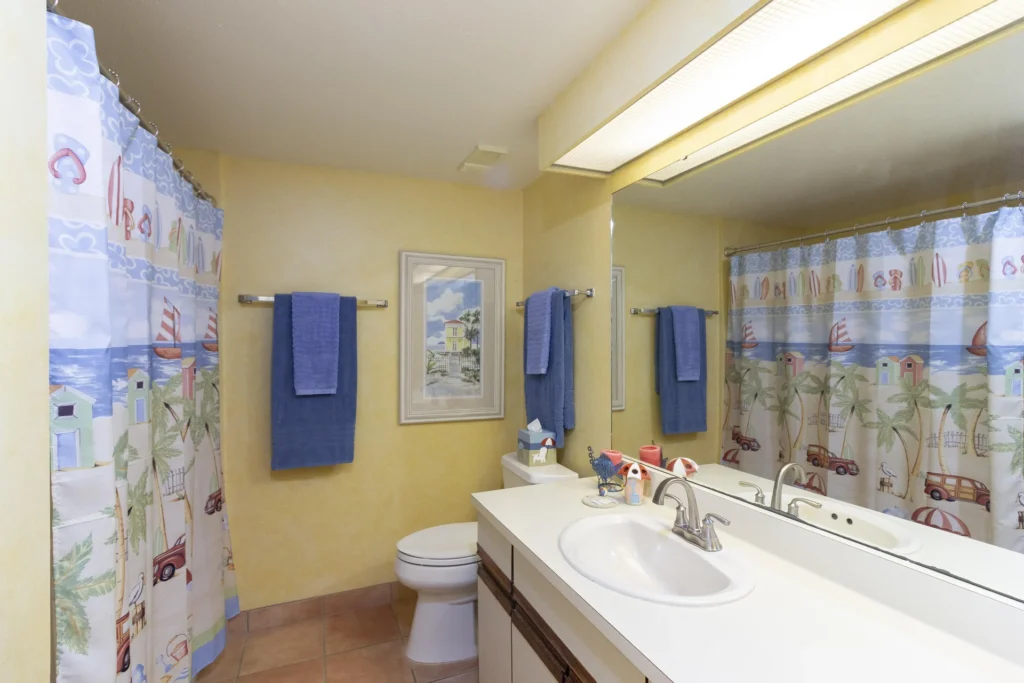 Bathroom with wide mirror, white countertop sink, patterned shower curtain, and blue towels on yellow walls.