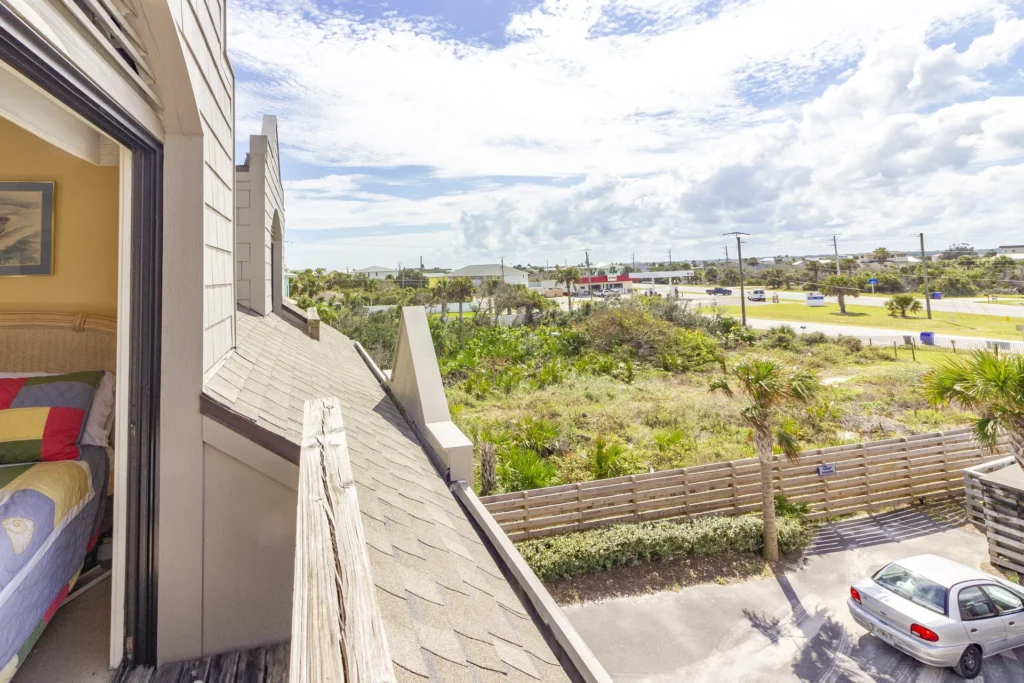 View from the balcony shows a sloped shingle roof, wooden railing, and greenery beyond the parking area.