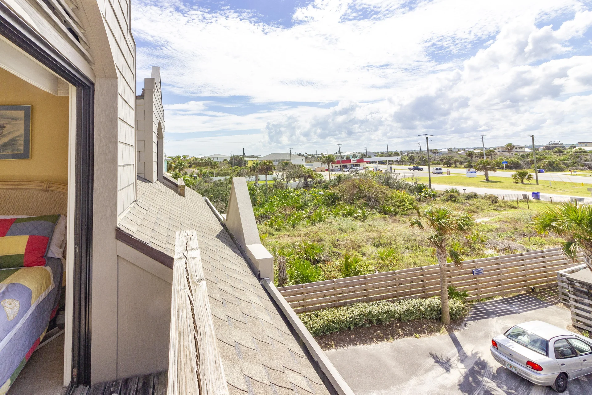 View from the balcony shows a sloped shingle roof, wooden railing, and greenery beyond the parking area.