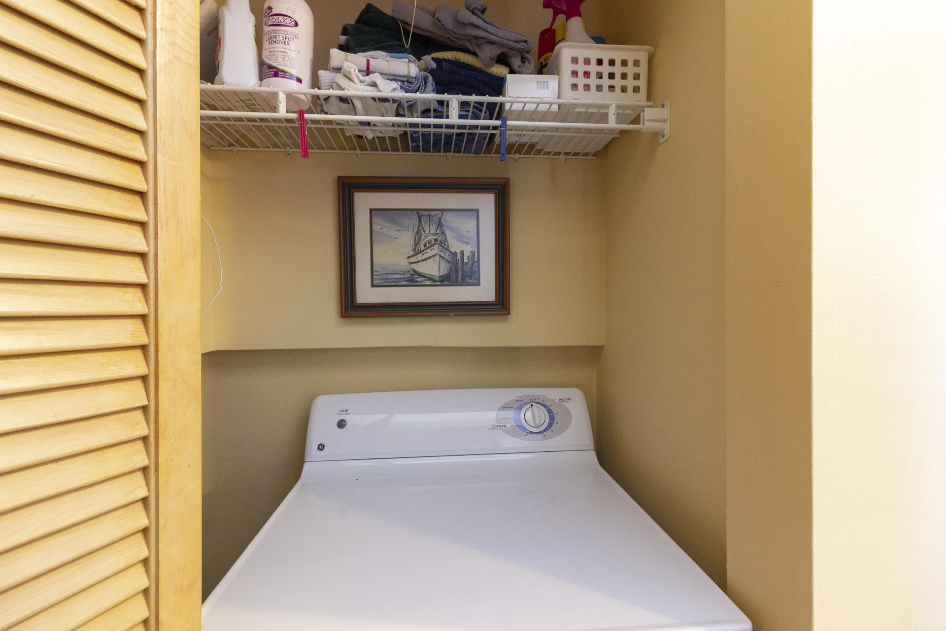 Laundry closet with white top-loading washer, beige walls, and a framed nautical print above the appliance.