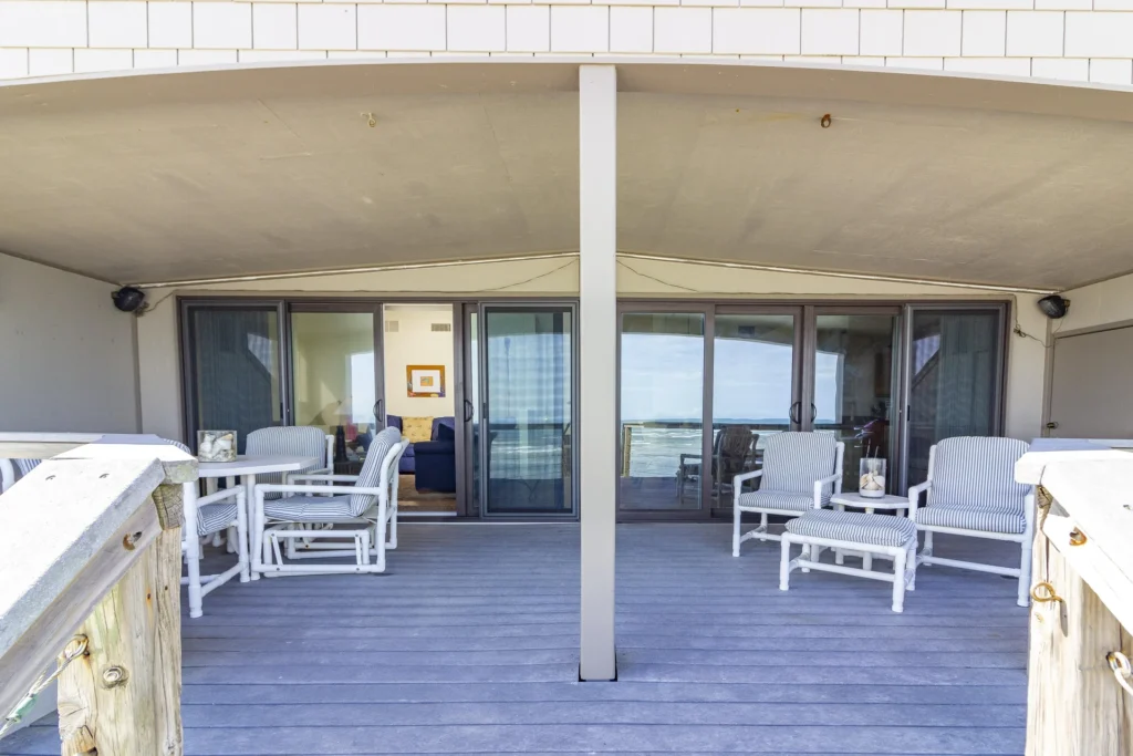 Oceanfront deck with gray composite flooring, white framed sliding glass doors, and cushioned striped seating groups.