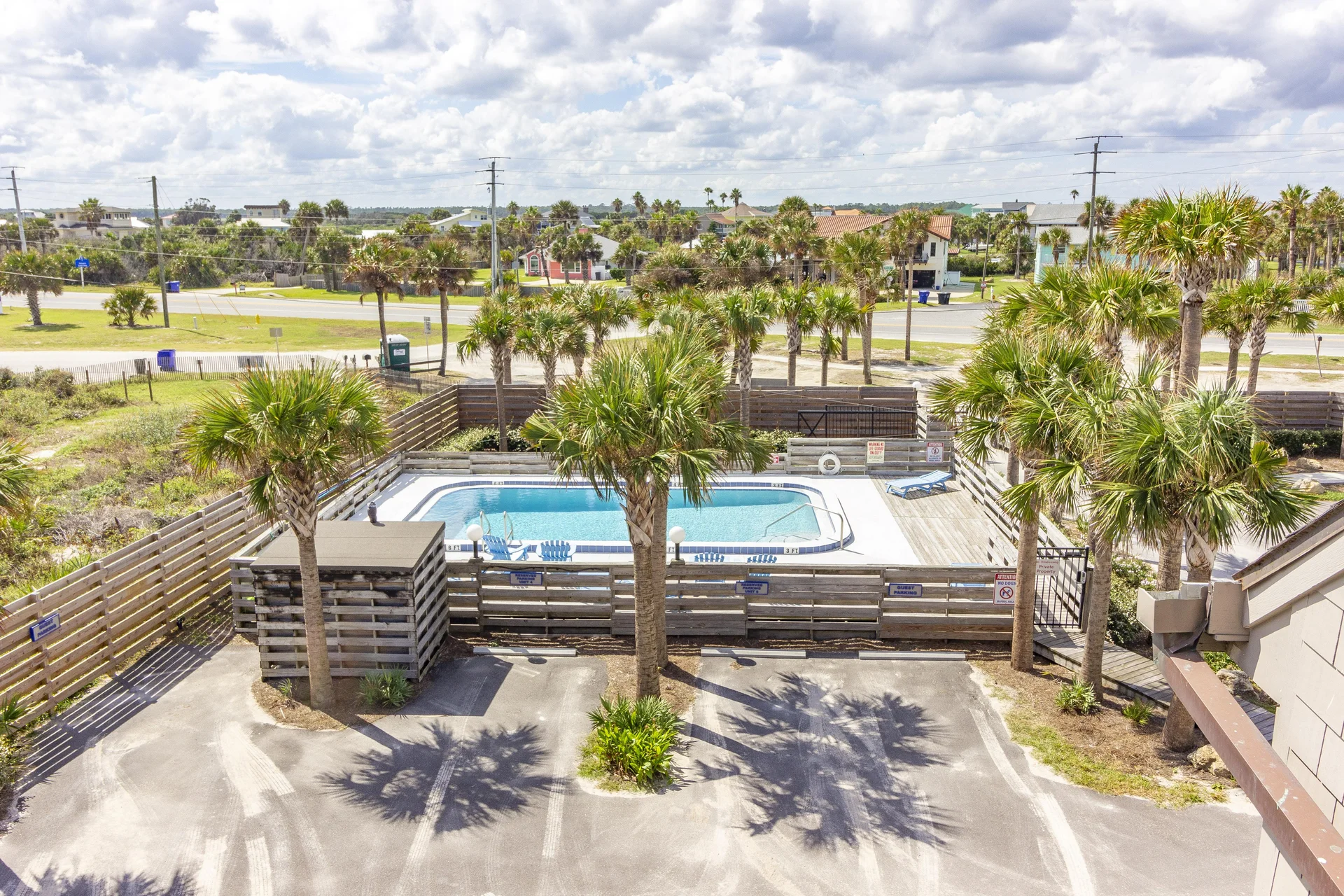 Gated outdoor pool area with white concrete deck, blue lounge chairs, surrounded by palm trees and wooden fencing.