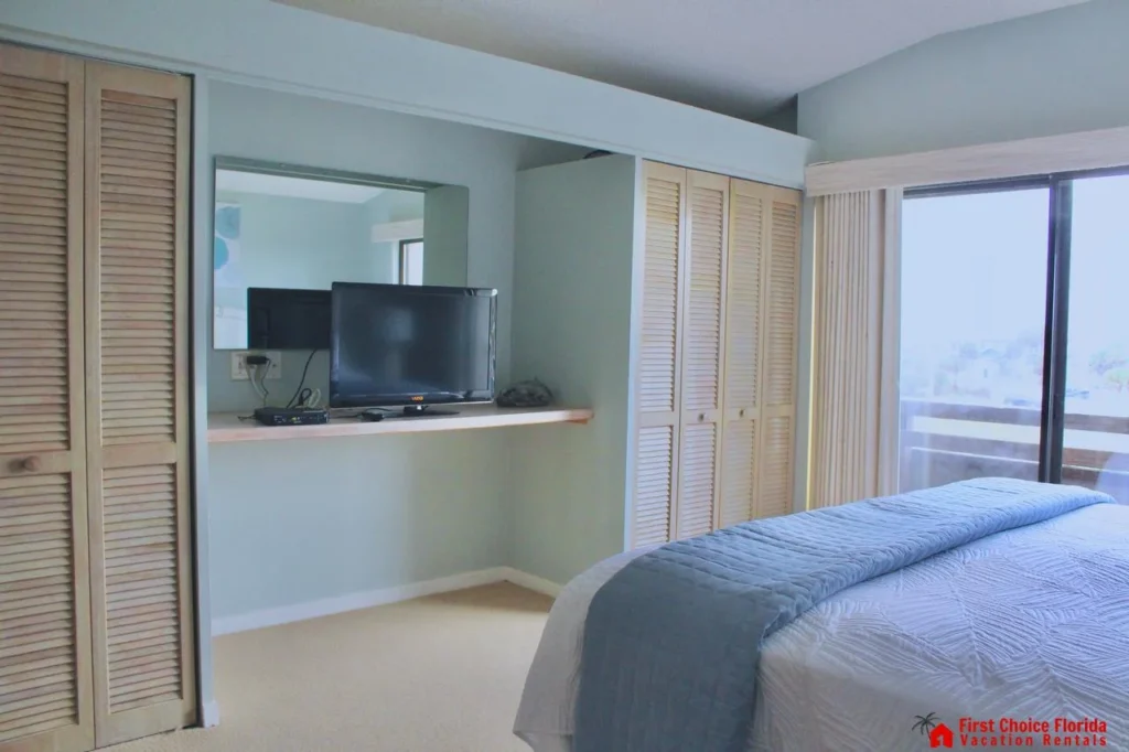 Mounted TV sits on a light wood shelf between two louvered closets in the bedroom with carpeted flooring.