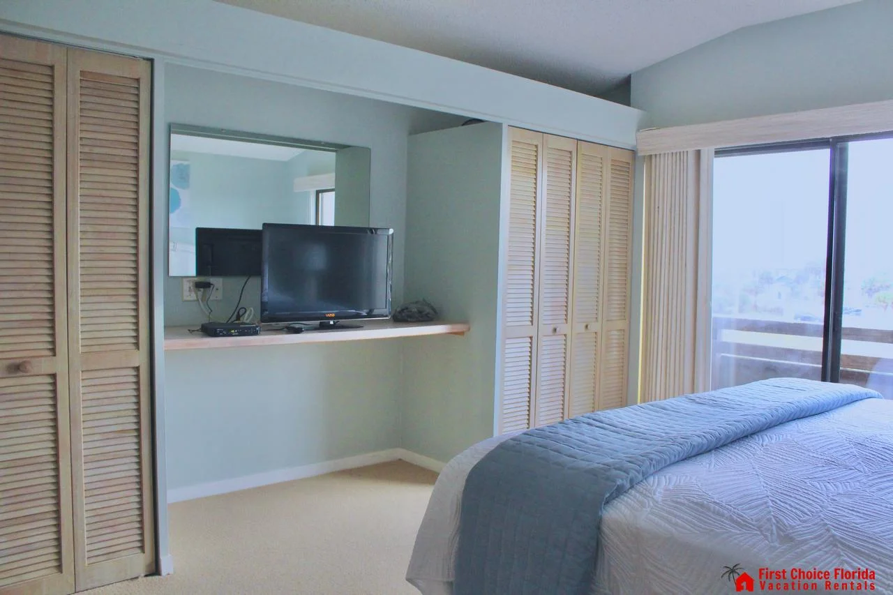 Mounted TV sits on a light wood shelf between two louvered closets in the bedroom with carpeted flooring.