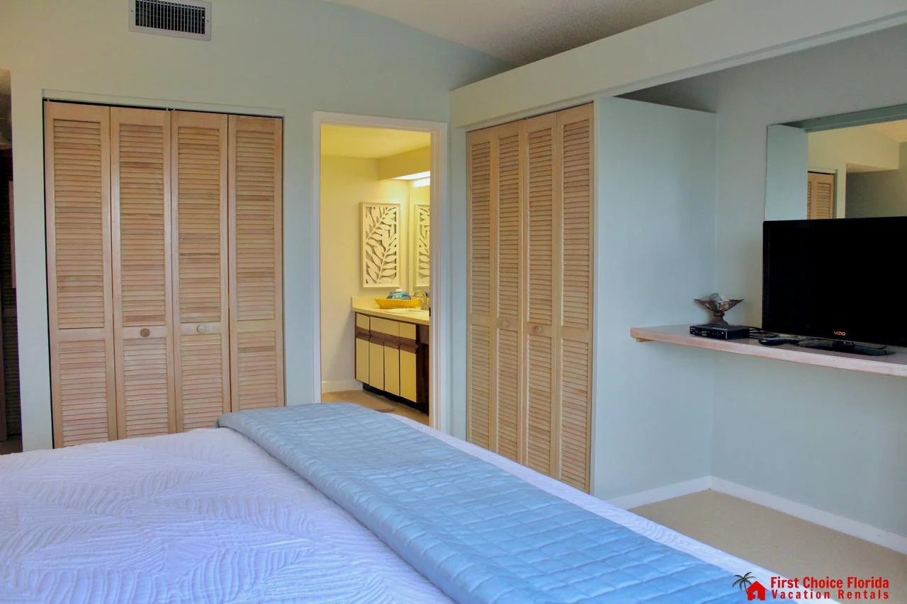 Bedroom with white quilted bedding, two louvered closets, and a wall-mounted TV above a wooden shelf.