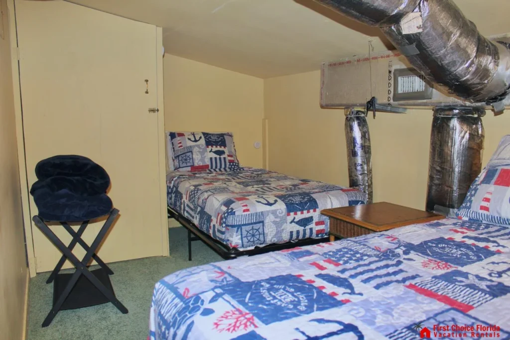 Small bedroom with twin beds dressed in nautical-themed quilts beneath exposed silver HVAC ducts.
