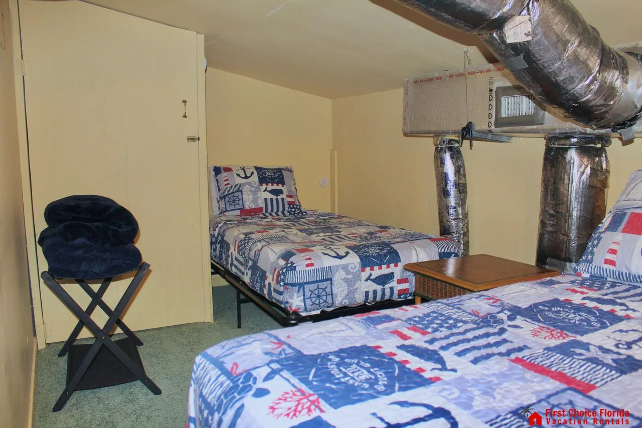 Small bedroom with twin beds dressed in nautical-themed quilts beneath exposed silver HVAC ducts.
