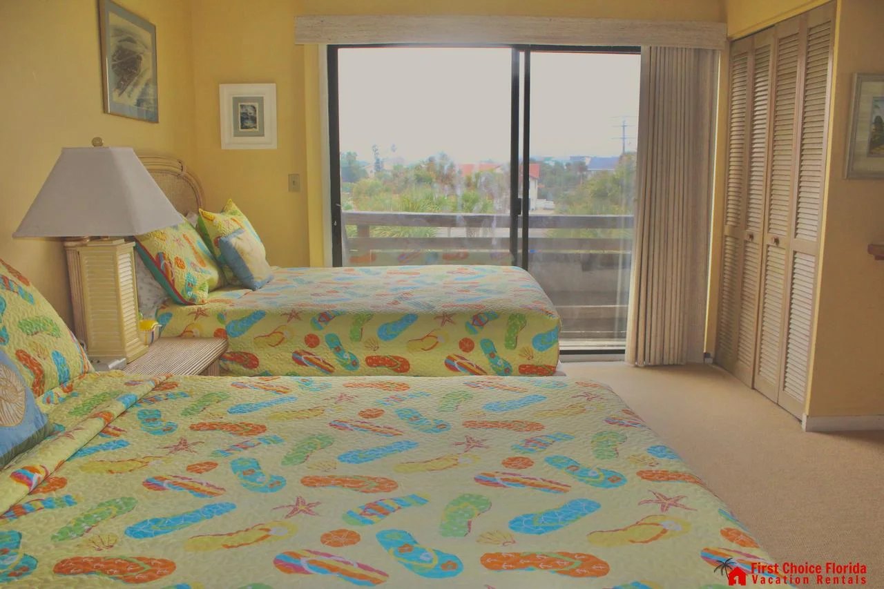 Two beds with colorful flip-flop patterned bedding face sliding glass doors opening to a balcony with outdoor views.