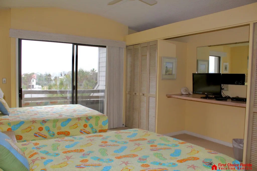 Two beds with colorful flip-flop patterned quilts face a wall-mounted TV and sliding glass door to the balcony.