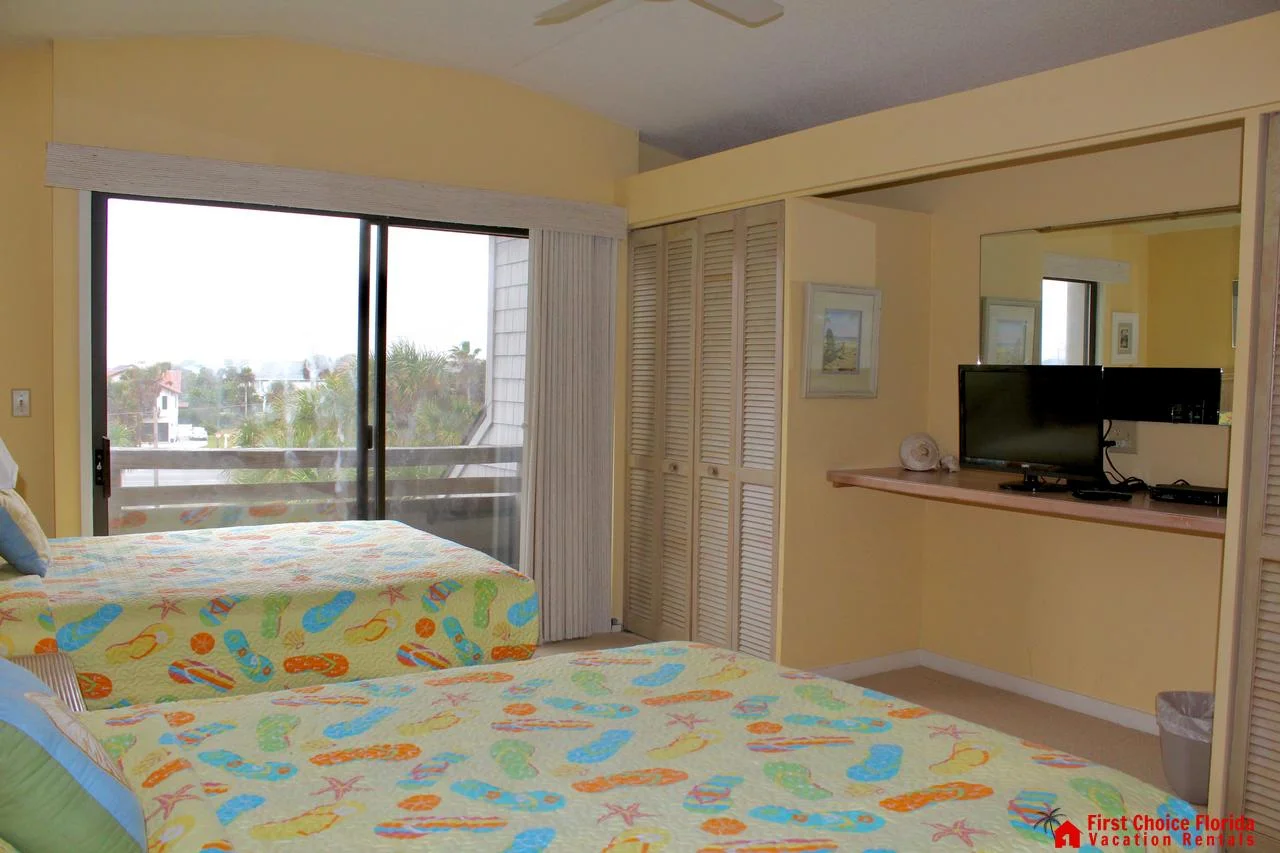Two beds with colorful flip-flop patterned quilts face a wall-mounted TV and sliding glass door to the balcony.