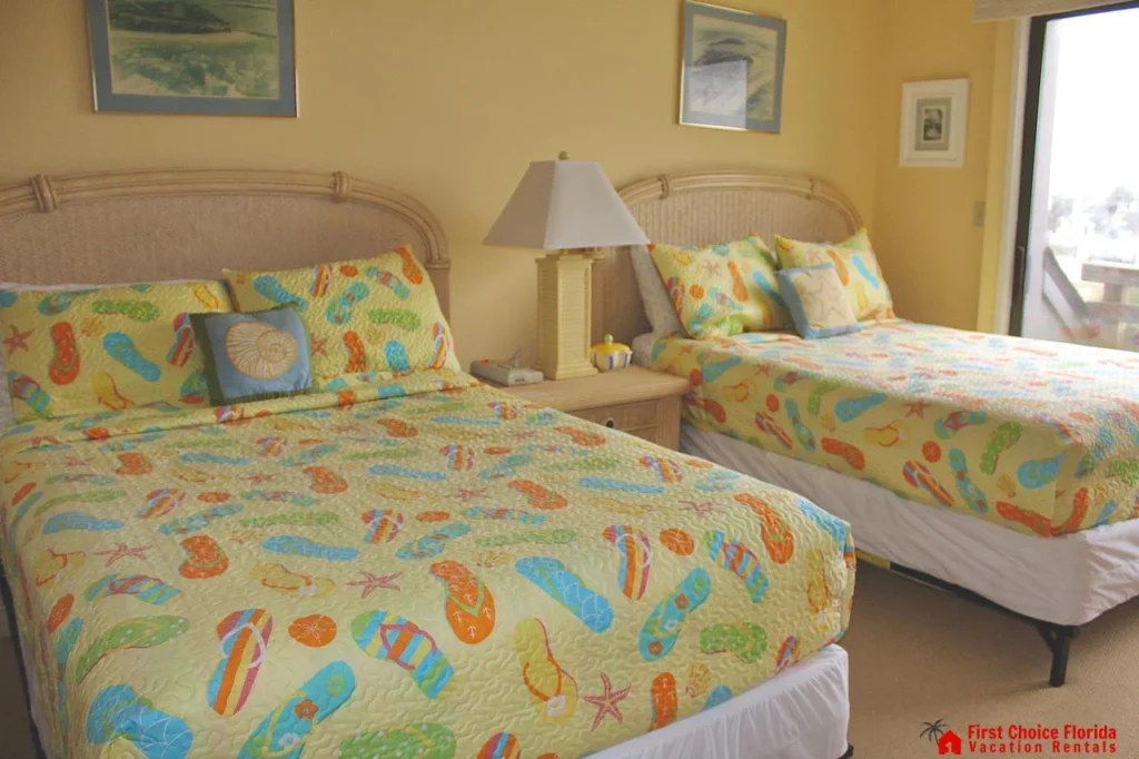 Two beds with wicker headboards and colorful flip-flop patterned bedding sit beneath framed beach art.