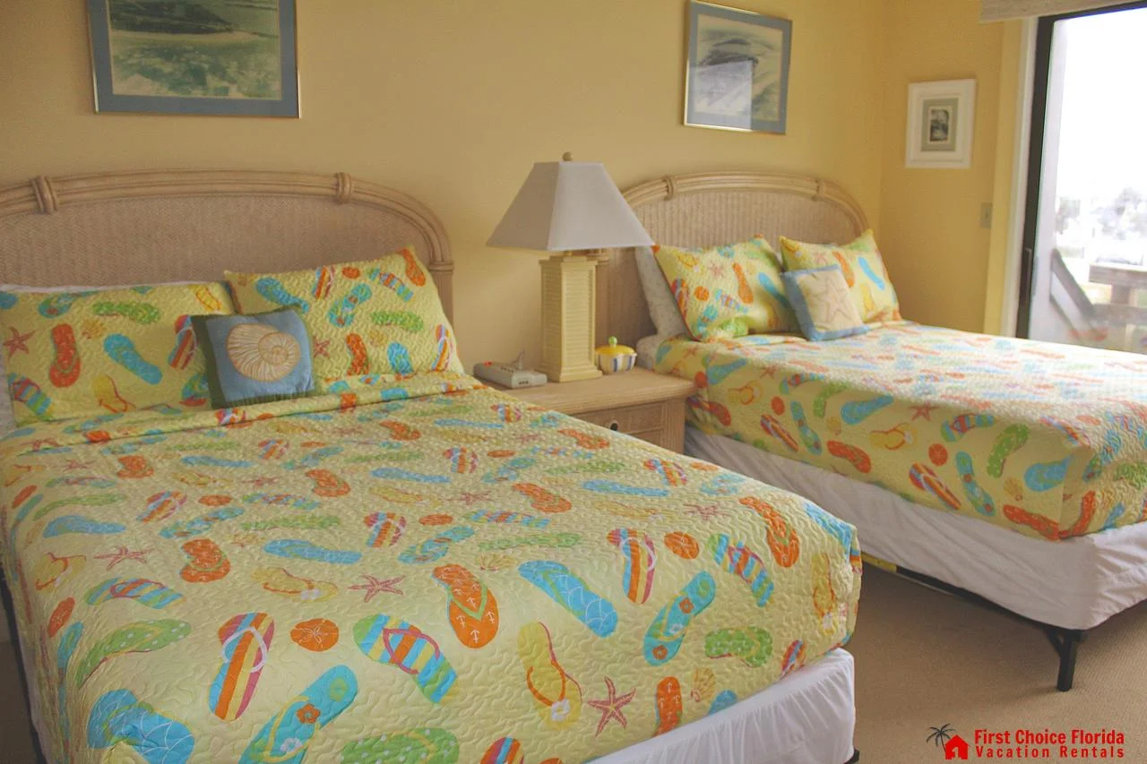 Two beds with wicker headboards and colorful flip-flop patterned bedding sit beneath framed beach art.