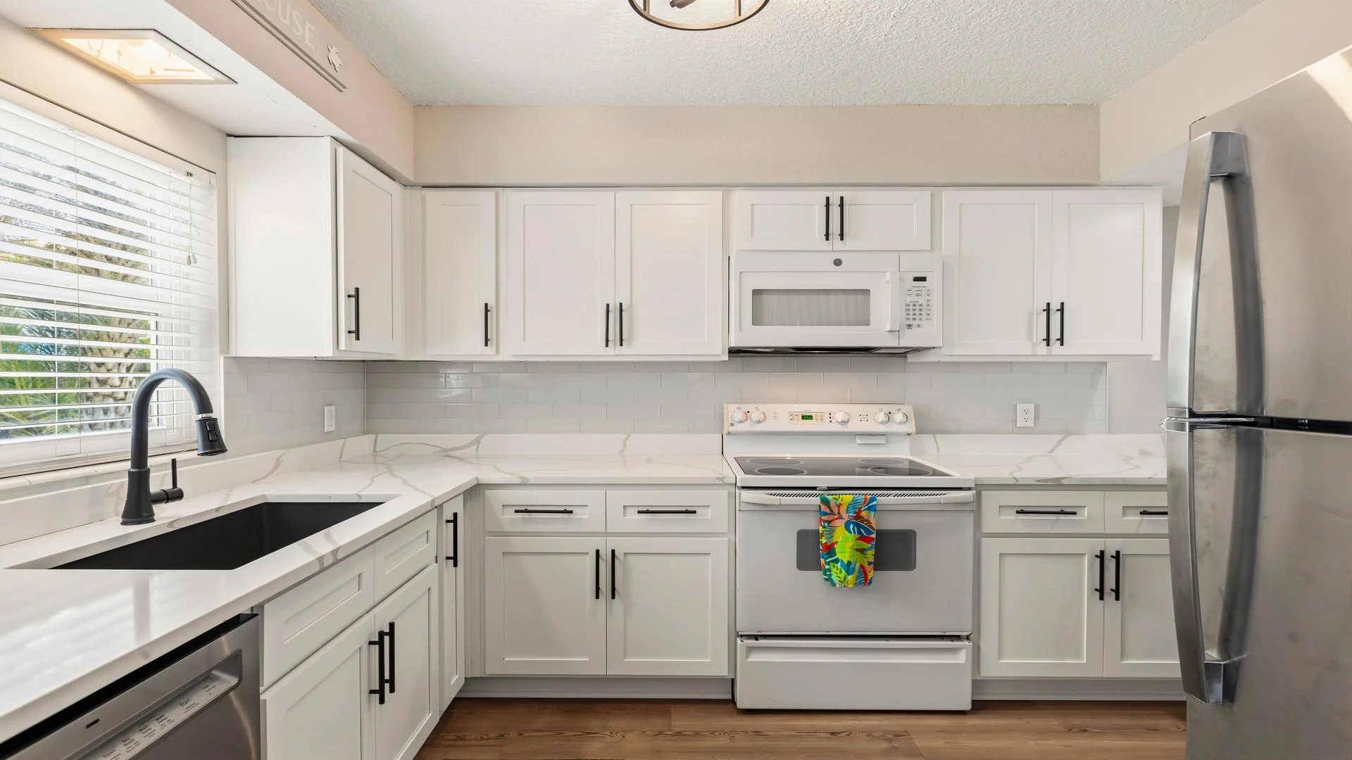 White cabinets with black hardware frame the marble-effect countertops and black sink under a large window.