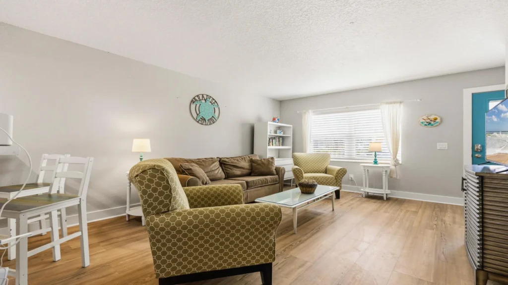 Living room with hardwood floors, patterned armchairs, brown sofa, glass coffee table, and a blue front door.
