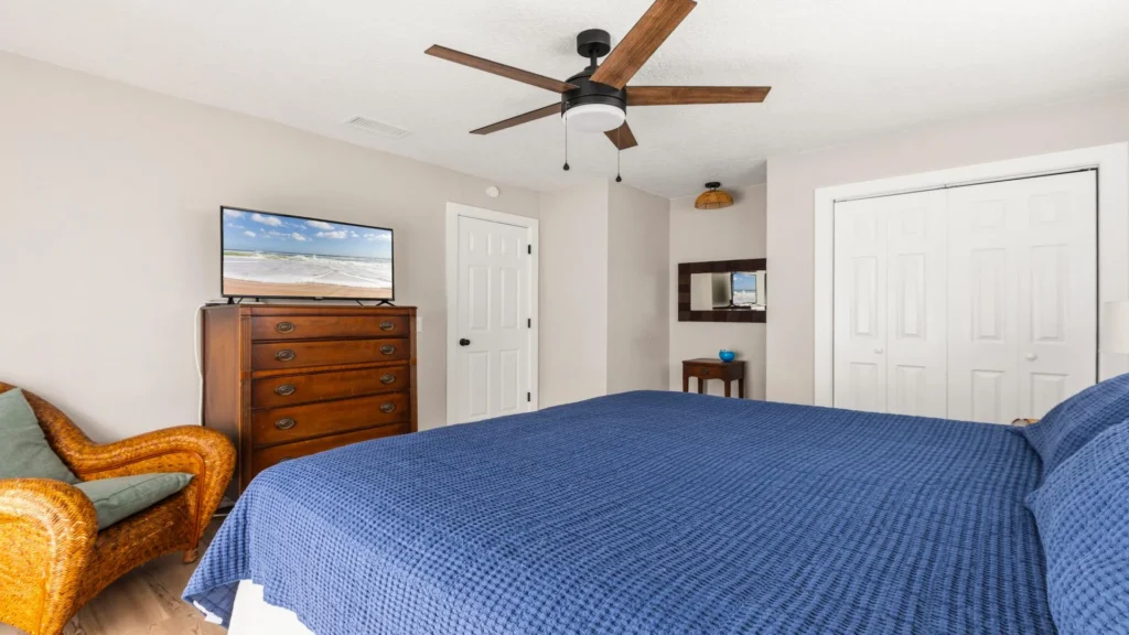 Primary bedroom with navy textured bedspread, wicker armchair, wooden dresser, and ceiling fan above.