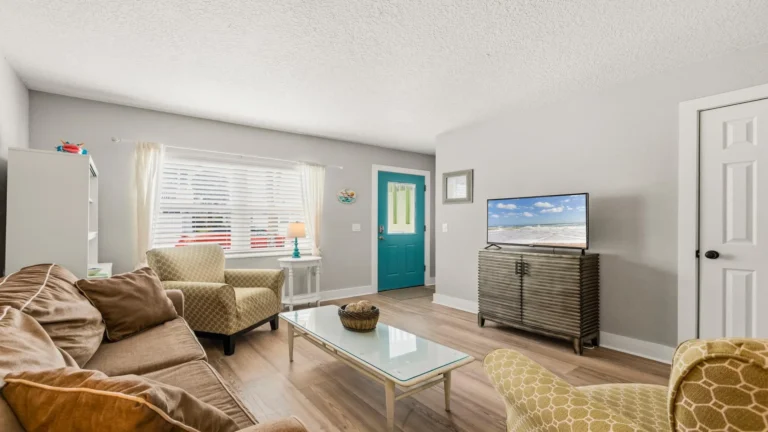 Living room with tan sofa, patterned armchairs, glass coffee table, wood flooring, and teal front door.