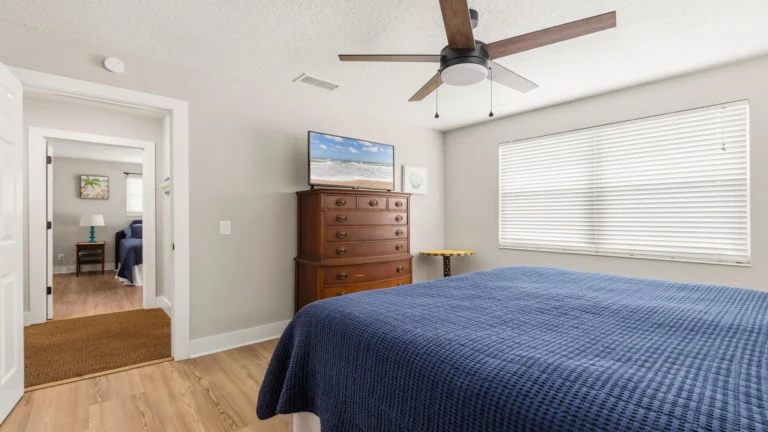 Primary bedroom with wood dresser under a wall-mounted TV, large window with white blinds, and ceiling fan.