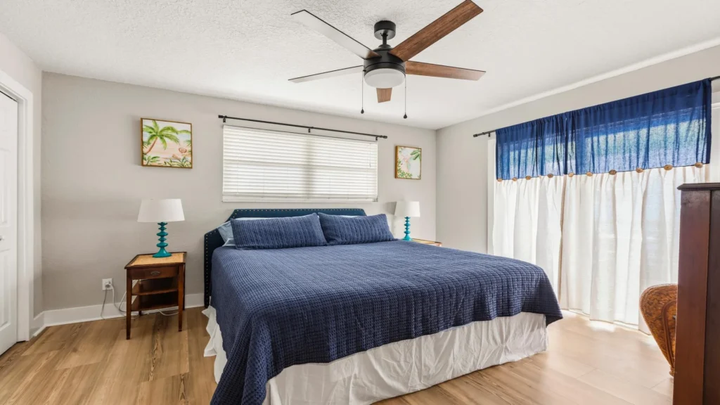 King bed with navy quilt and matching pillows beneath a wide window with white blinds and coastal artwork.