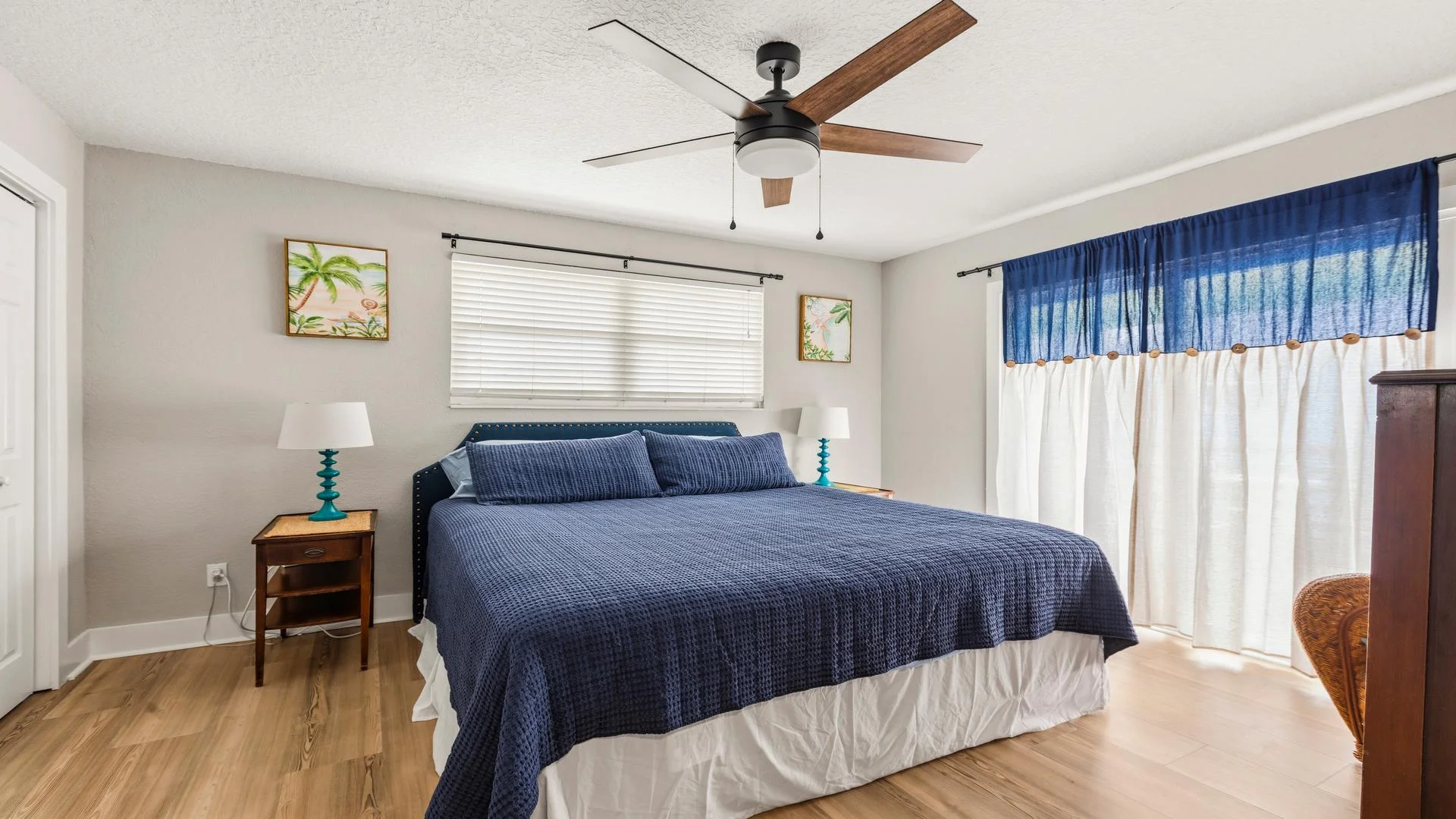 King bed with navy quilt and matching pillows beneath a wide window with white blinds and coastal artwork.