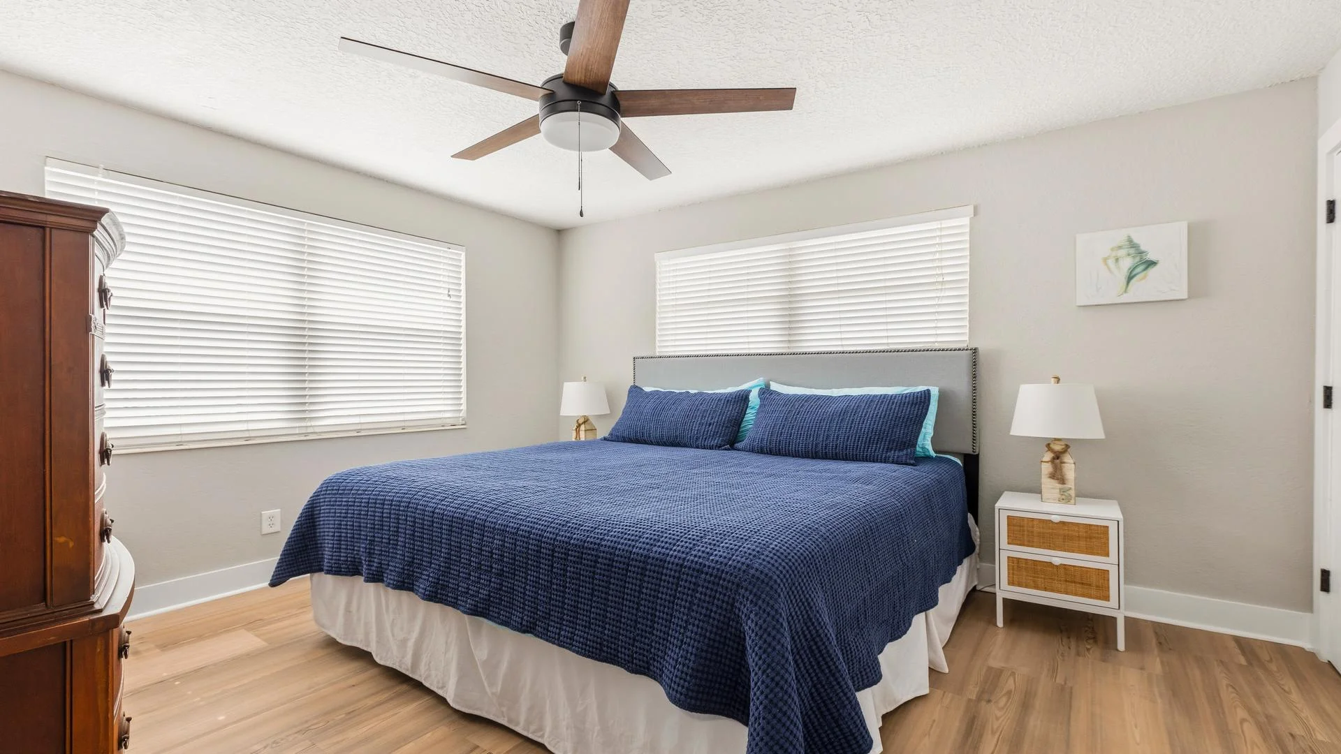 Primary bedroom with wood flooring, blue textured bedding, twin nightstands, and a ceiling fan with wood blades.