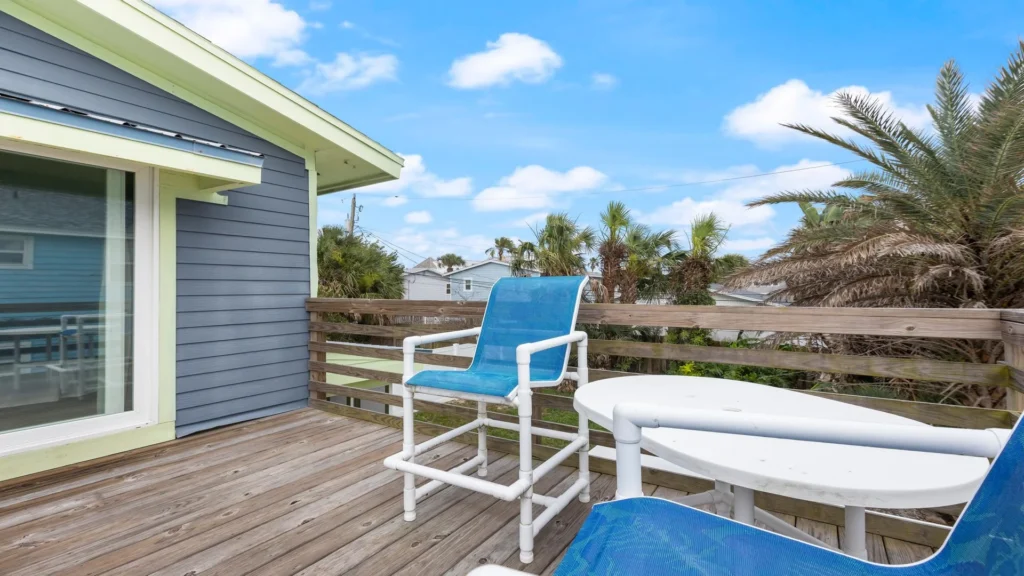Second-floor balcony with weathered wood deck, white outdoor table, blue mesh chairs, and palm tree views.