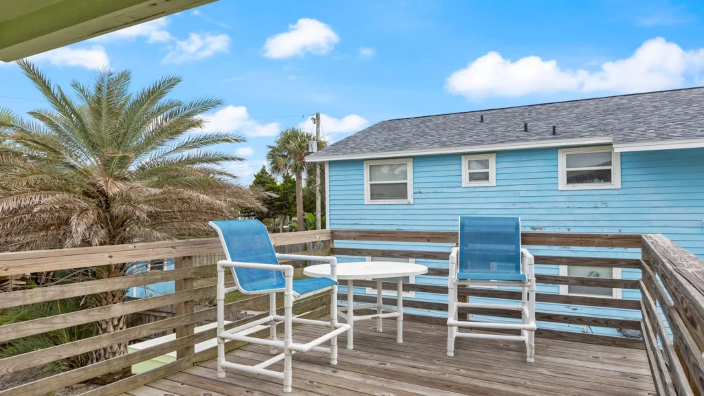 Wood deck balcony with white and blue sling chairs and round white table overlooking palm trees.