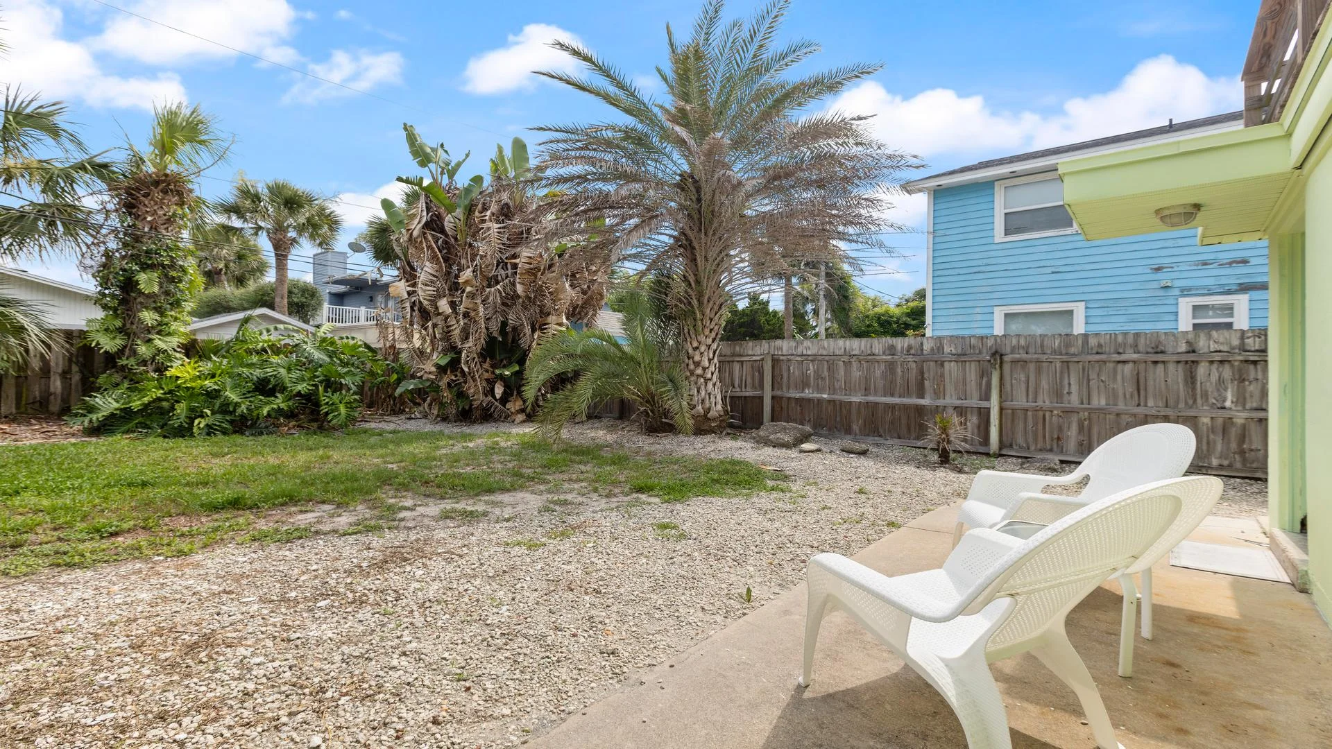 Patio with lightweight white chairs overlooks a gravel yard enclosed by a wooden fence and tropical plants.