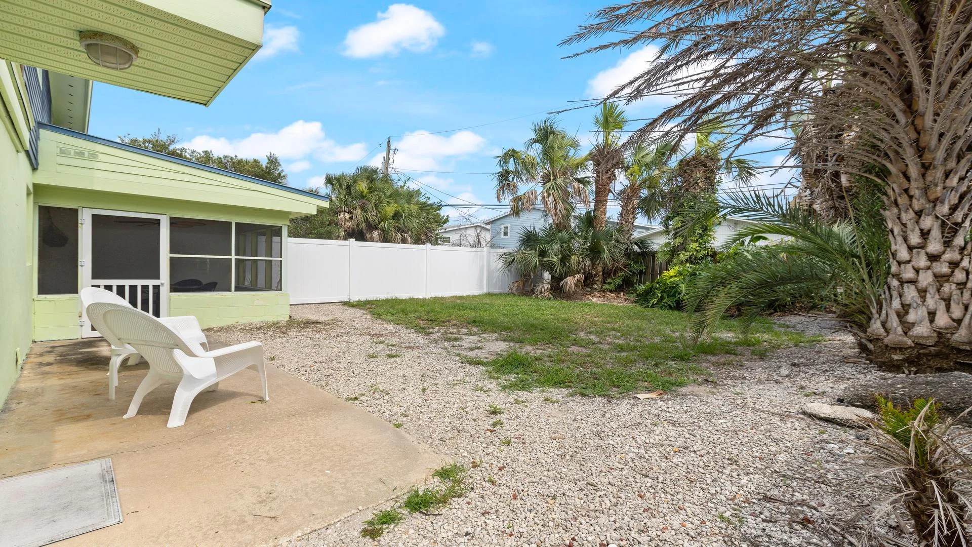 Backyard patio with two white plastic chairs, gravel ground, palm trees, and a white privacy fence.