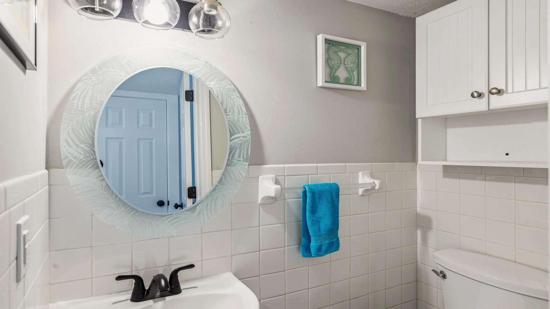 Half bathroom features white tile wainscoting, a round etched glass mirror, and white cabinetry above the toilet.
