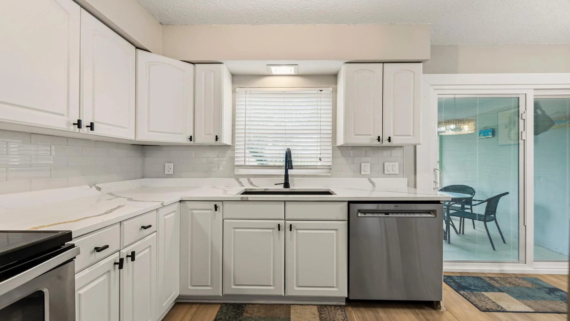 White cabinetry and marble-effect countertops surround a black faucet and stainless dishwasher near sliding glass doors.