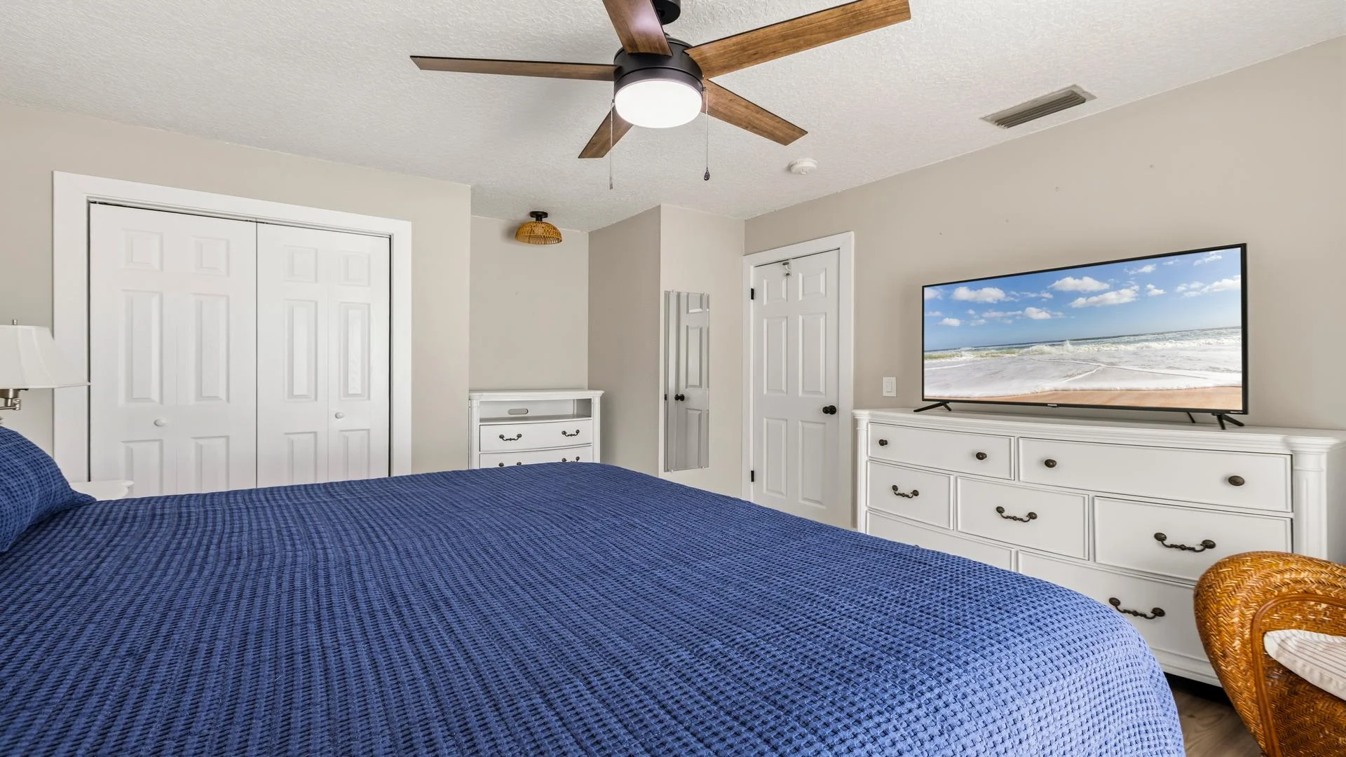 Bedroom with navy textured bedspread, white dressers, wicker chair, ceiling fan, and wall-mounted TV.