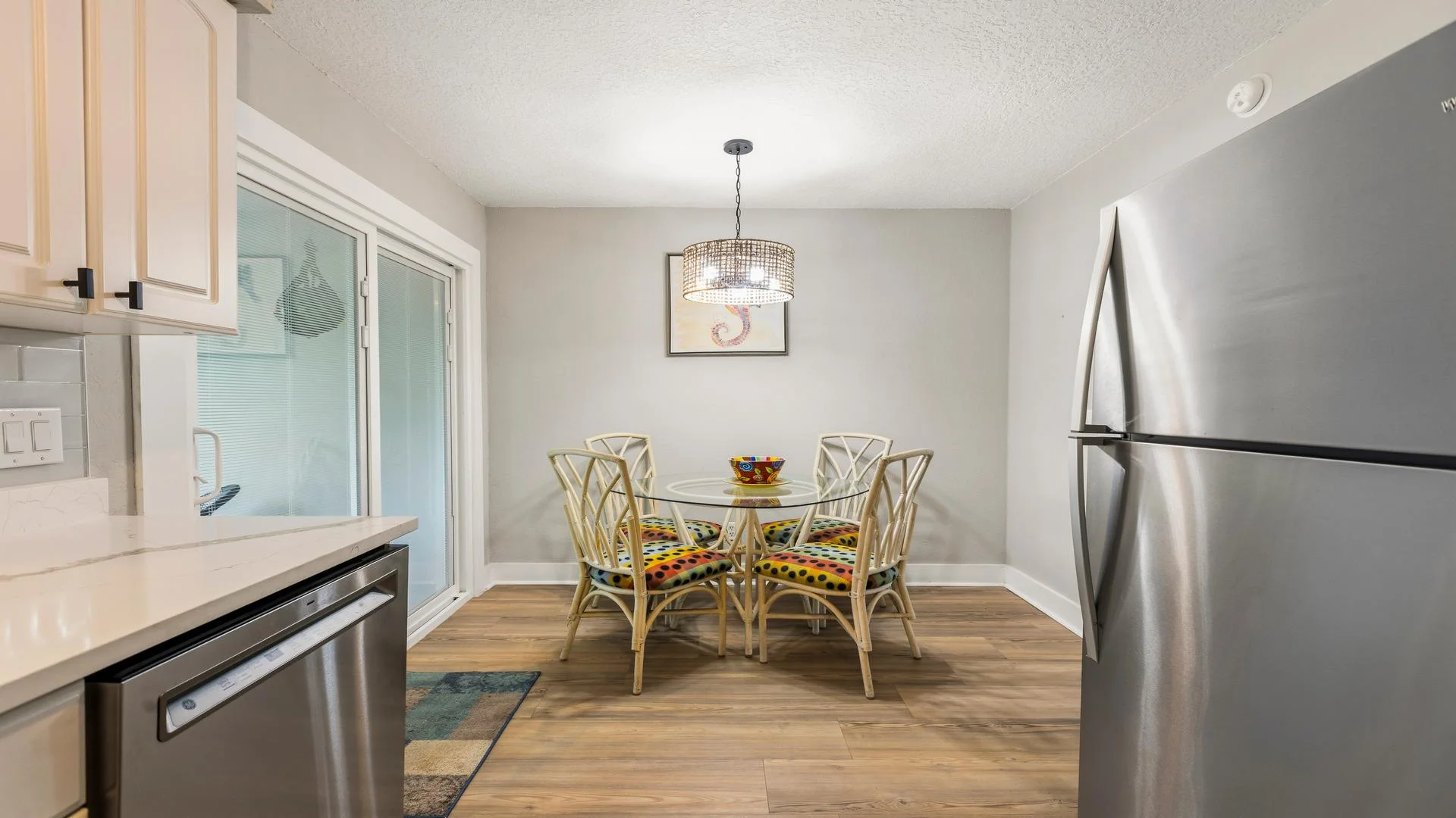 Dining area with glass-top table, colorful cushioned rattan chairs, pendant light, and wood-look flooring.