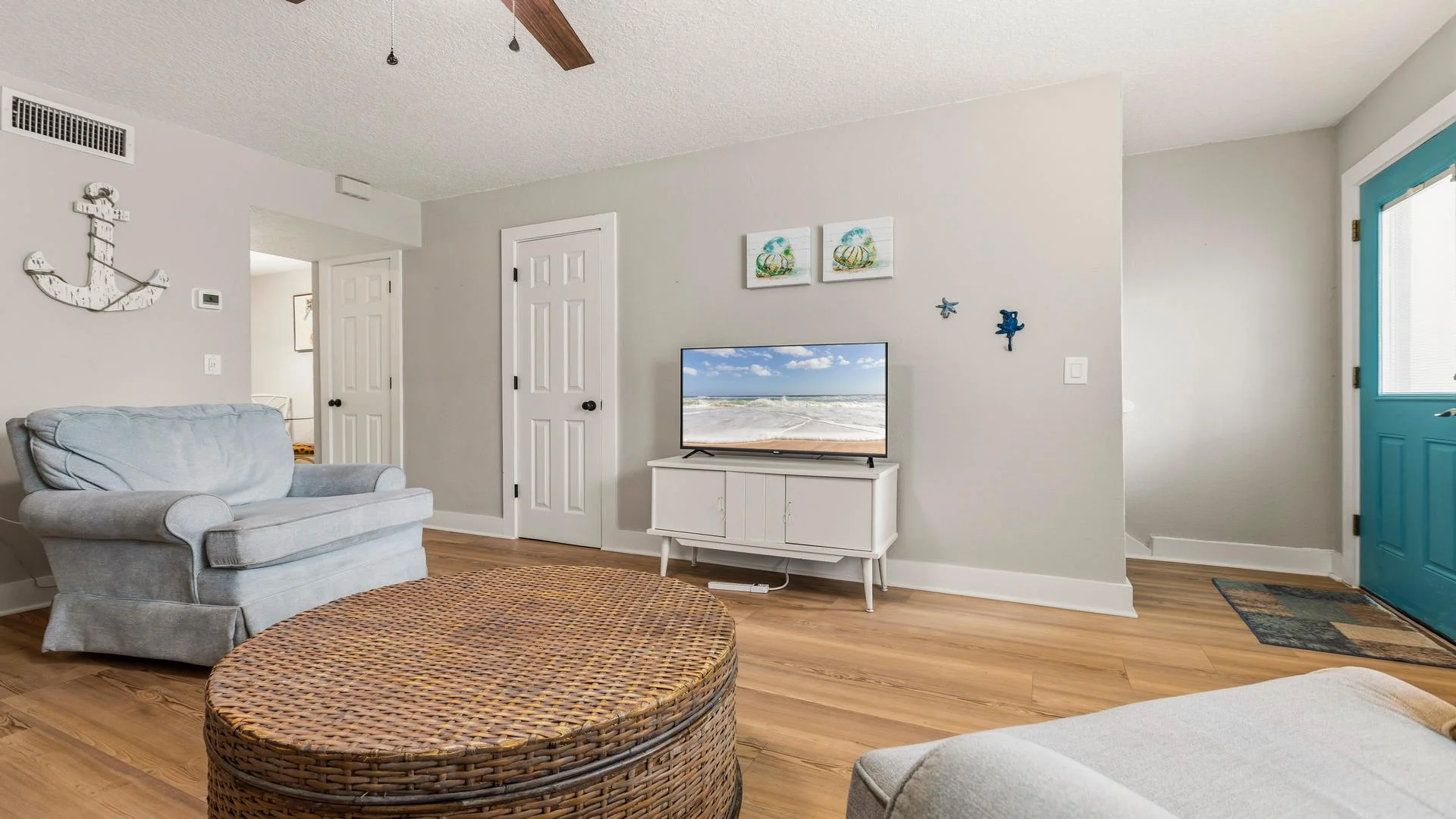 Living room with light blue armchairs, a woven round coffee table, wood-look flooring, and a TV on a white console.