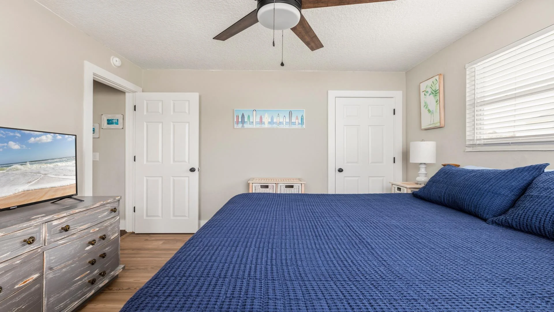 Primary bedroom with navy textured bedding, weathered gray dresser, ceiling fan, and coastal-themed wall art.