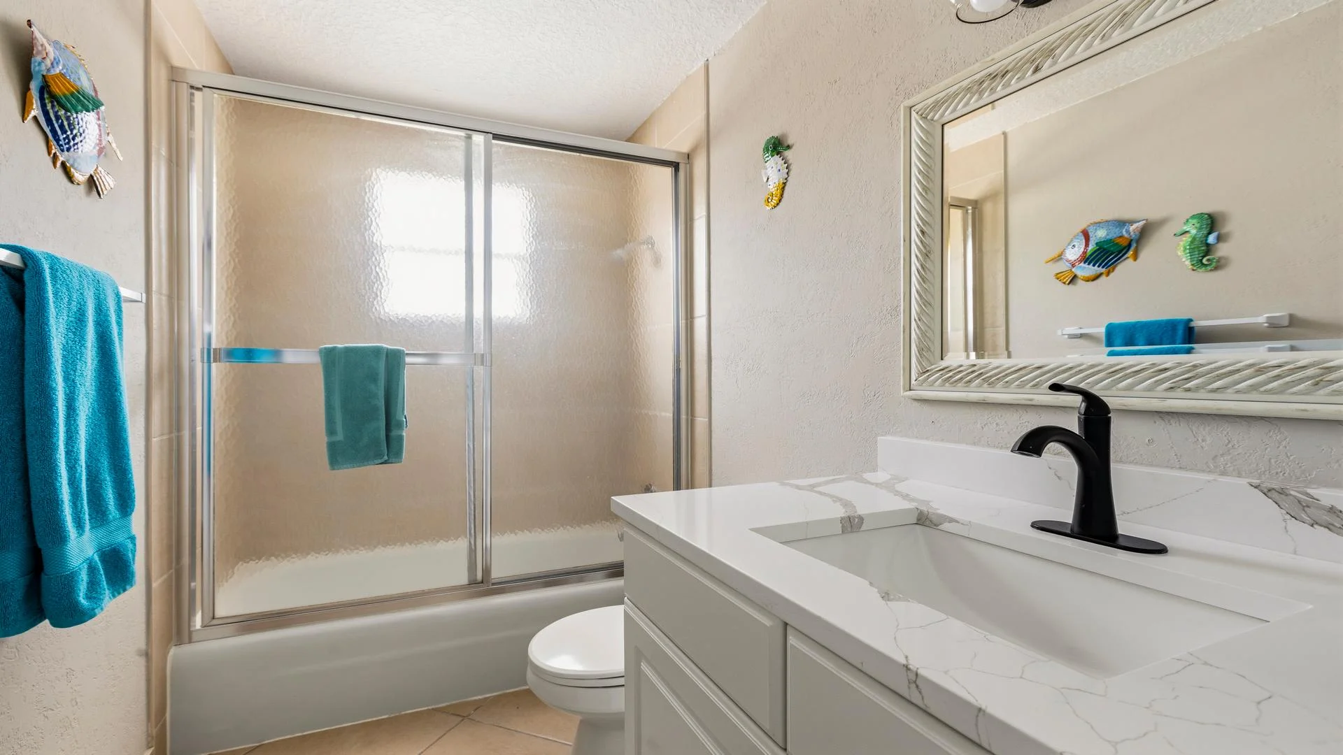 Bathroom with marble-style quartz countertop, black faucet, textured glass shower doors, and ocean-themed wall decor.