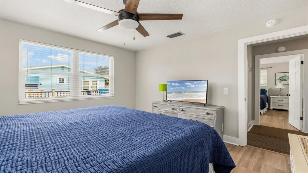 Blue quilt covers the bed beneath a ceiling fan in the main bedroom with a gray wood dresser and window views.