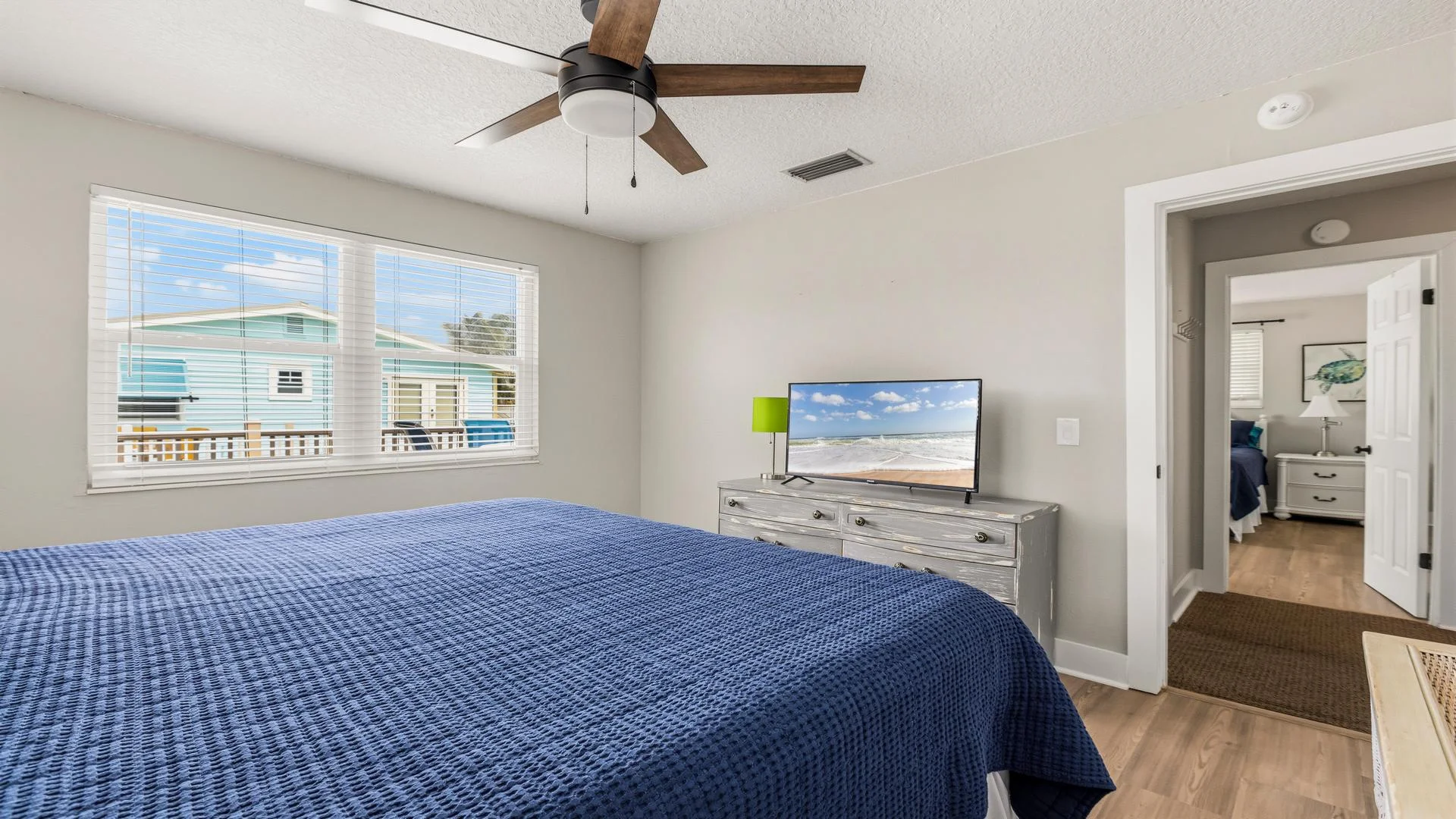 Blue quilt covers the bed beneath a ceiling fan in the main bedroom with a gray wood dresser and window views.