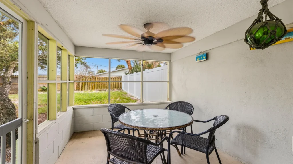 Screened porch with concrete floor, black wicker chairs around a glass-top table, and ceiling fan blades in motion.