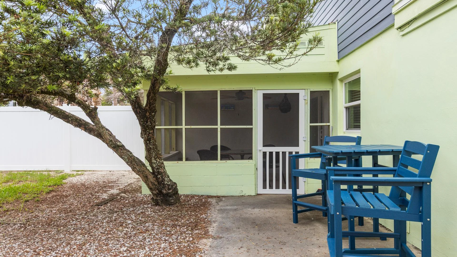Concrete patio with blue wooden chairs and table beside a screened porch and pale green exterior walls.